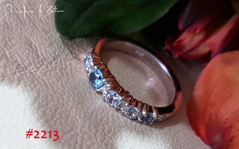Anillo plateado #2213 con topacio azul central y línea de cristales brillantes sobre fondo claro con elementos florales rojos