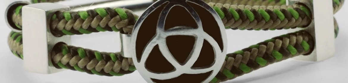 Primer plano de una pulsera hecha a mano con dos hebras de cordón trenzado en tonos tierra y verde. Las hebras están unidas por un broche rectangular de metal plateado y un adorno circular que presenta un diseño de triquetra recortado, revelando un fondo oscuro contrastante. El enfoque nítido en la pulsera resalta la artesanía y los materiales utilizados.