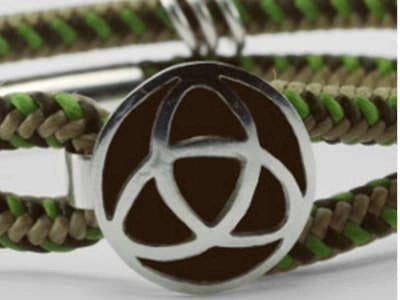 Pulsera trenzada de doble hebra con adorno de trisquel Primer plano de una pulsera hecha a mano con dos hebras de cordón trenzado en tonos tierra y verde. Las hebras están unidas por un broche rectangular de metal plateado y un adorno circular que presenta un diseño de triquetra recortado, revelando un fondo oscuro contrastante. El enfoque nítido en la pulsera resalta la artesanía y los materiales utilizados.