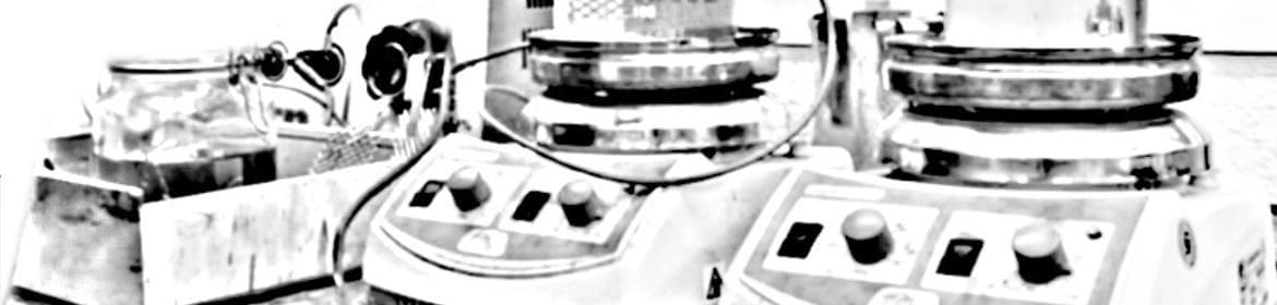 Una imagen en escala de grises de varios equipos de laboratorio, posiblemente incluyendo placas calefactoras, un agitador magnético y otros instrumentos electrónicos. La imagen parece estar tomada en un entorno de laboratorio y los cables que conectan los equipos son visibles. La imagen monocromática y la calidad ligeramente granulada sugieren que podría ser una fotografía antigua o una copia escaneada.