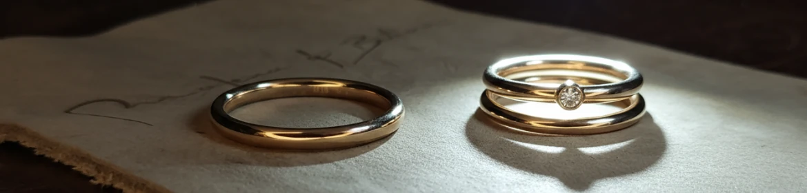  Dos anillos de boda de oro sobre una superficie clara con escritura borrosa. A la izquierda, un anillo liso de oro. A la derecha, un conjunto de dos anillos de oro, uno con un pequeño diamante central, ambos iluminados con un brillo.