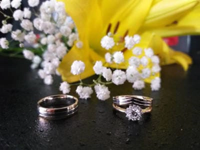 Primer plano de dos anillos de boda sobre una superficie oscura, junto a flores blancas y un lirio amarillo, representando la unión matrimonial y la celebración del 'sí quiero