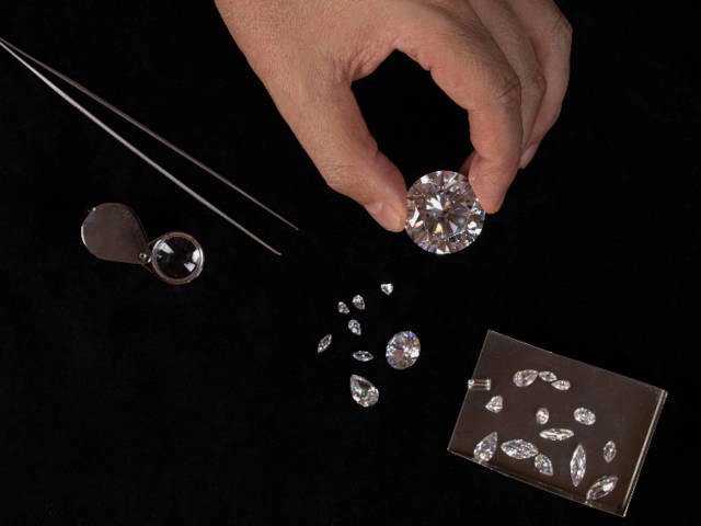 Clasificación y examen de diamantes Diamantes naturales vs diamantes de laboratorio La imagen muestra una mano sosteniendo un diamante grande con pinzas sobre un fondo negro. Se ven varios otros diamantes de diferentes tamaños y cortes dispersos sobre la superficie, junto con una lupa y pinzas, herramientas que se utilizan para examinar y clasificar los diamantes. Esto sugiere un entorno profesional, como el de un gemólogo o un clasificador de diamantes, que evalúa la calidad y las características de las piedras.