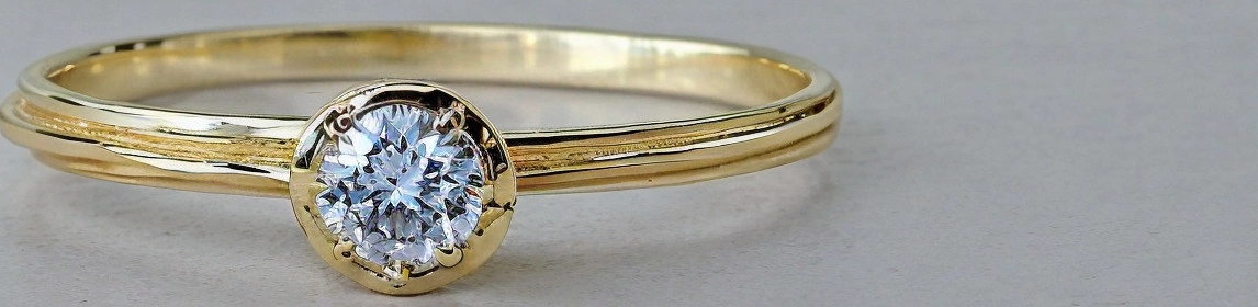 Un anillo de oro con un diamante solitario engastado en bisel se muestra sobre un fondo liso y de color claro. El anillo tiene una banda de oro simple con un diseño de doble línea que envuelve el dedo. El diamante es redondo y brillante, y está asegurado por un engaste de bisel circular que lo rodea por completo. La imagen está enfocada en el anillo, destacando el brillo del diamante y el pulido del oro.
