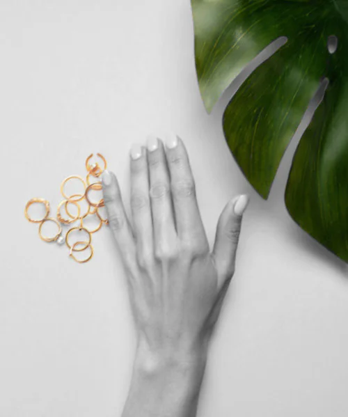 Una mano femenina con manicura blanca y sencilla se muestra sobre un fondo blanco. Un montón de anillos de oro finos y delicados se encuentra cerca de la mano. Una hoja verde tropical se encuentra en la esquina superior derecha de la imagen. La imagen es una composición elegante y minimalista.